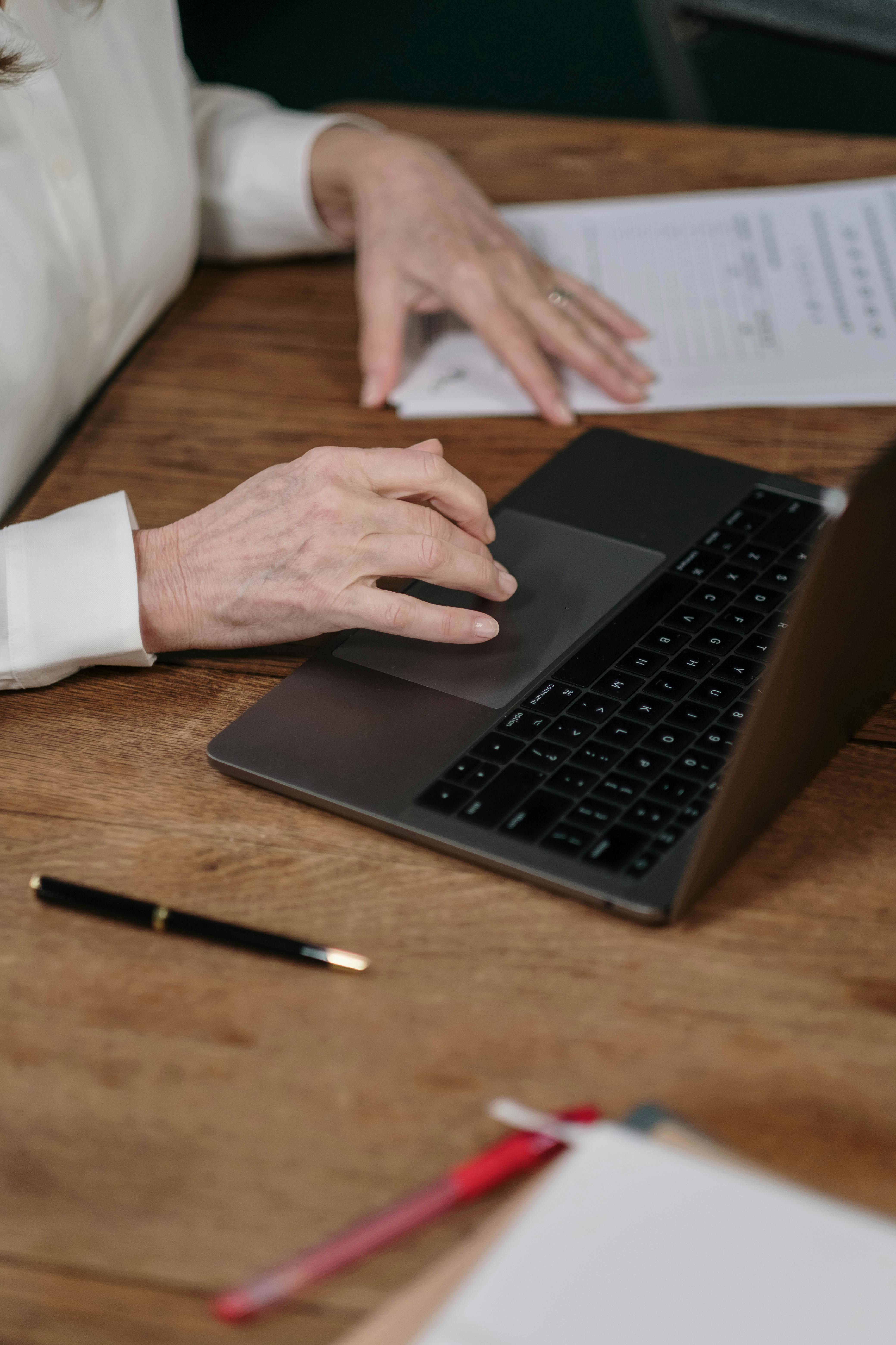 From above of crop anonymous mature female entrepreneur in formal clothes reading documents and working on laptop at table in office